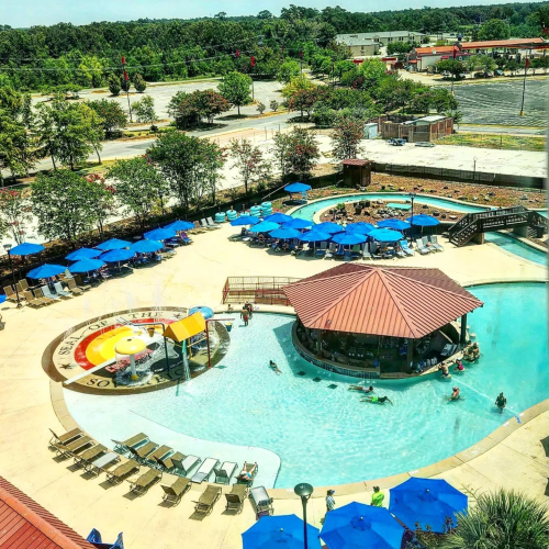 Aerial view of a resort pool area with blue umbrellas, lounge chairs, and a water slide surrounded by greenery.