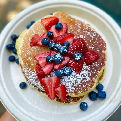 A stack of pancakes topped with fresh strawberries, blueberries, and a dusting of powdered sugar on a paper plate.