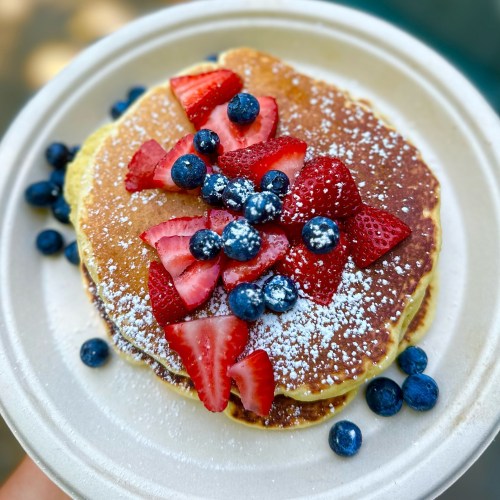 A stack of pancakes topped with fresh strawberries, blueberries, and a dusting of powdered sugar on a paper plate.