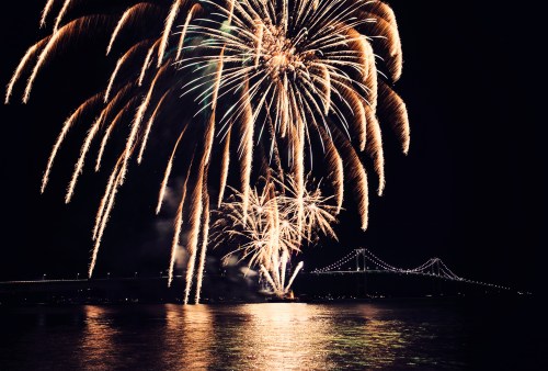 Colorful fireworks illuminate the night sky above a bridge, reflecting on the water below.