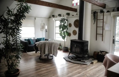 Cozy living room with a wood stove, plants, and a comfortable couch, featuring wooden beams and warm decor.