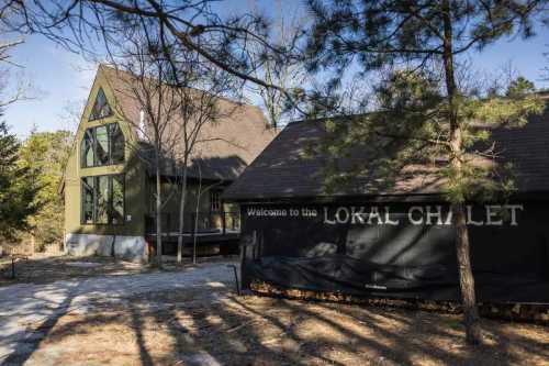 A cozy chalet with a green exterior and a sign reading "Welcome to the Lokal Chalet," surrounded by trees.