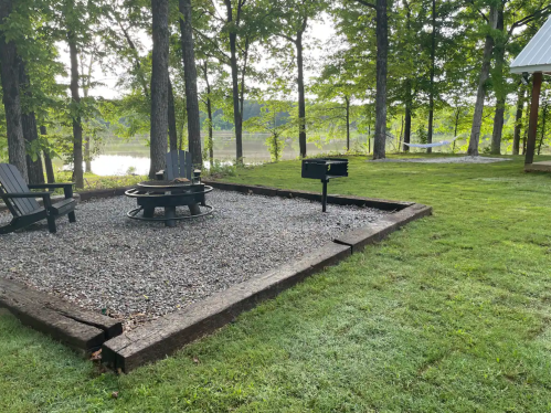 A serene outdoor space with a fire pit, grill, and seating area surrounded by trees and a view of a lake.