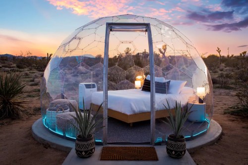 A transparent dome with a bed inside, surrounded by desert landscape and a colorful sunset sky.