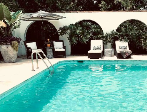 A serene pool area with turquoise water, lounge chairs, an umbrella, and lush greenery in the background.