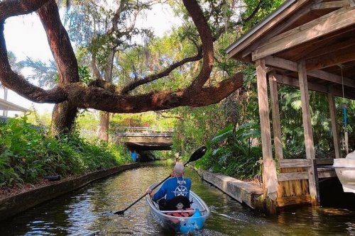 A person in a kayak paddles through a lush, green waterway lined with trees and a wooden structure.