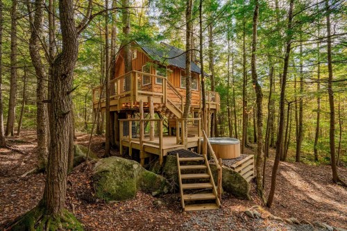 A wooden treehouse nestled among trees, featuring a deck, stairs, and a hot tub on a rocky foundation.