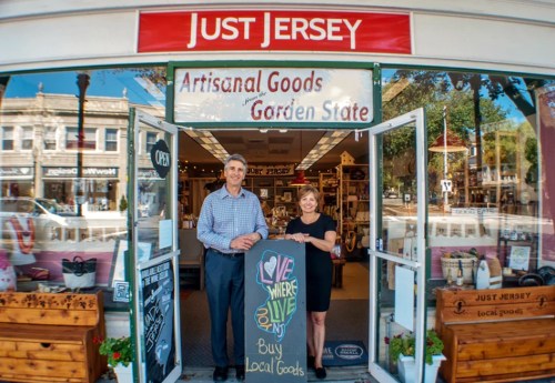 A man and woman stand at the entrance of "Just Jersey," a shop selling artisanal goods, with a welcoming sign.