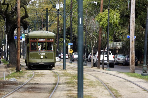 A green streetcar travels along tracks in a tree-lined urban area, with cars parked nearby and street signs visible.