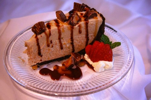 A slice of peanut butter cheesecake topped with chocolate drizzle, mini peanut butter cups, and a strawberry on the side.