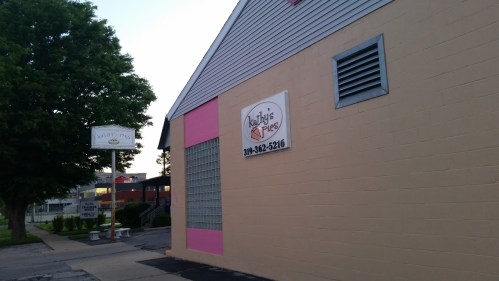 Exterior view of a building with a sign for "Kathy's Pies" and a phone number, surrounded by trees and a sidewalk.