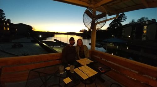 A couple sits at a table on a patio, enjoying a sunset view over a lake with menus and drinks in front of them.