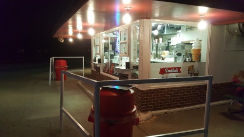 A nighttime view of a small ice cream stand with bright lights and a service window, surrounded by red trash bins.