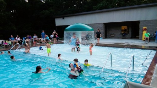 A lively pool scene with children playing, a water feature, and adults relaxing on the deck in the background.