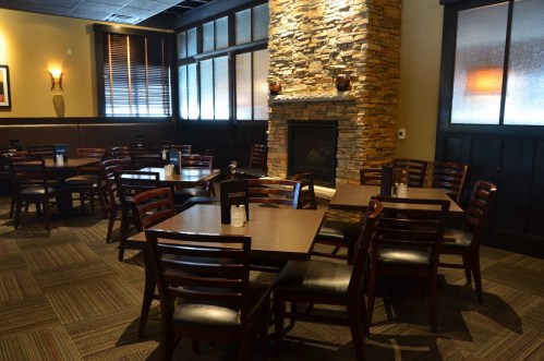 Cozy restaurant interior with wooden tables, chairs, and a stone fireplace, featuring warm lighting and a welcoming atmosphere.