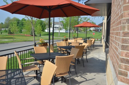 Outdoor café seating with wicker chairs and orange umbrellas, overlooking a green landscape and a quiet street.