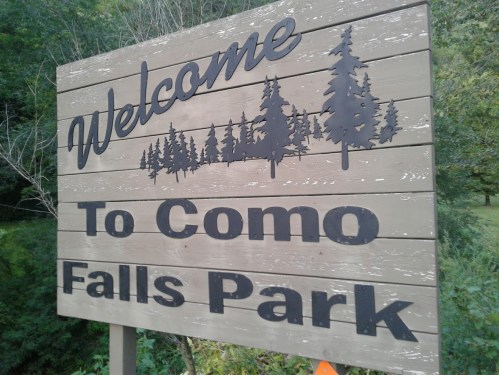 Welcome sign for Como Falls Park, featuring trees and a natural setting.