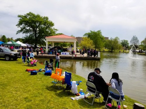 People enjoying a sunny day by a pond, with tents and a gazebo in the background, and children playing nearby.