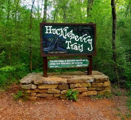 Sign for Huckleberry Trail surrounded by greenery, featuring a wooden structure and a quote by Mark Twain.