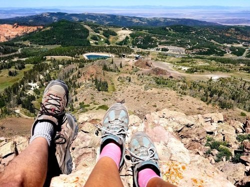 Two pairs of feet in hiking shoes resting on a rocky ledge, overlooking a scenic valley and distant mountains.