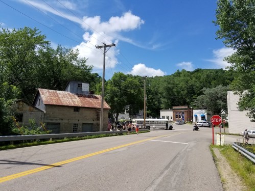 A rural road scene with a stop sign, old buildings, and trees under a blue sky with clouds.