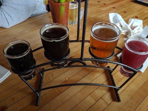 A flight of four craft beers in small glasses on a metal holder, set on a wooden table.