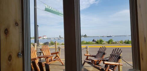 View from a wooden deck with chairs, overlooking a calm waterfront and boats in the distance under a clear sky.