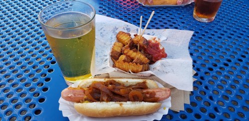 A hot dog with onions, a serving of crinkle-cut fries, and a glass of beer on a blue table.