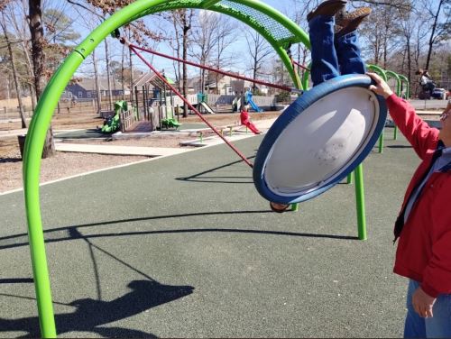 A child swings upside down on a circular swing while an adult holds the swing's edge at a playground.