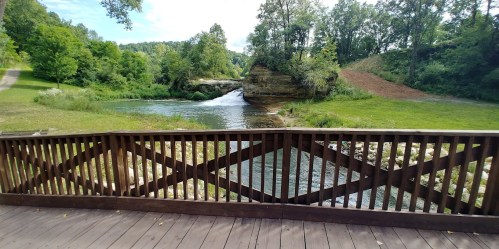 A scenic view of a river with a small waterfall, surrounded by lush greenery and trees, seen from a wooden deck.