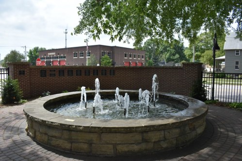 A circular stone fountain with water jets, surrounded by greenery and a brick wall, in a small town setting.