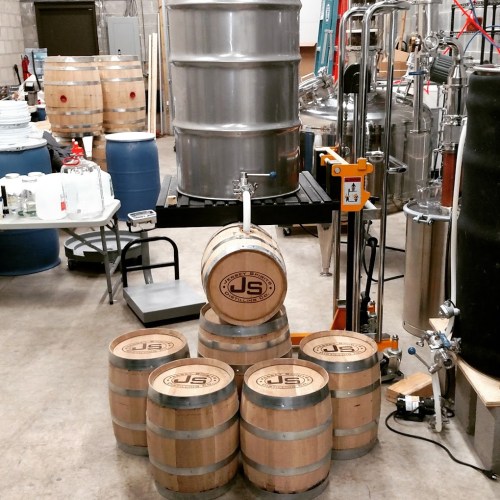 A distillery setup featuring wooden barrels, a stainless steel tank, and various brewing equipment in a spacious room.