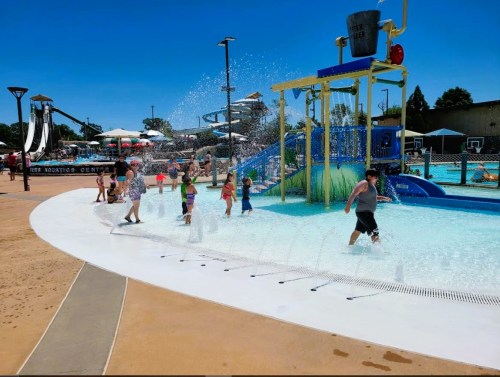 Children play in a water park, splashing under a large water bucket with slides and umbrellas in the background.