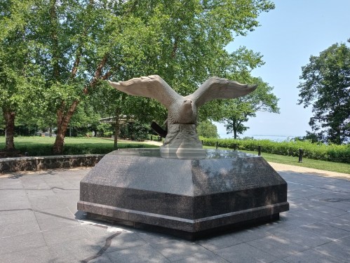 A stone eagle sculpture with outstretched wings, surrounded by trees in a park setting.
