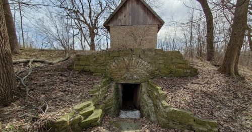 An old stone structure with a wooden roof, surrounded by trees and overgrown foliage, leading to a dark entrance.