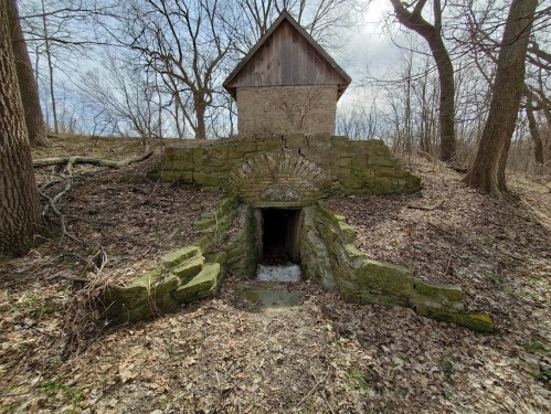 An old stone structure with a wooden roof, surrounded by trees and bare ground, leading to a dark entrance.