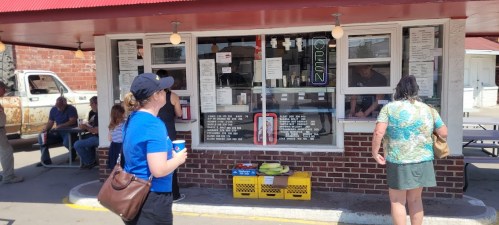 A woman in a blue shirt orders at a food stand with a menu, while others wait nearby. Sunny day, casual atmosphere.