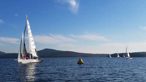 Sailboats on a calm lake with mountains in the background and a yellow buoy in the foreground. Clear blue sky.