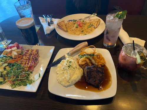 A table with three plates of food: a steak with mashed potatoes, pasta, and a salad, alongside drinks.