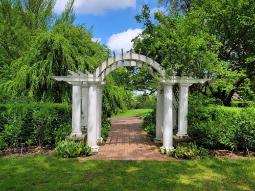 A white archway surrounded by lush greenery and trees, leading to a pathway in a serene garden setting.
