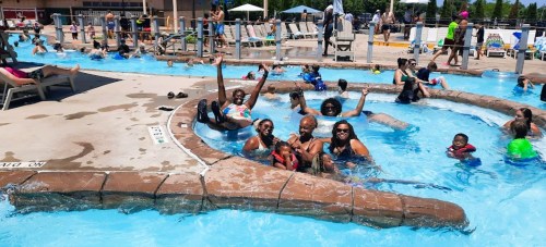 A lively pool scene with families and friends enjoying the water, some waving and smiling, under a sunny sky.