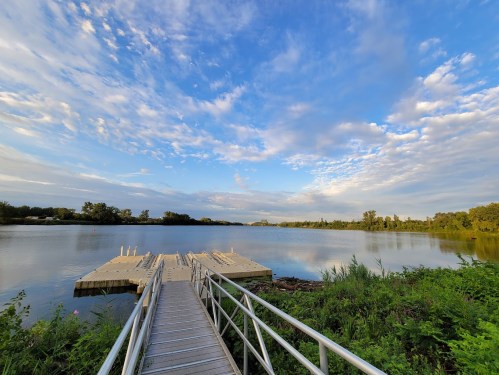 A wooden dock extends into a calm river, surrounded by greenery and under a blue sky with scattered clouds.