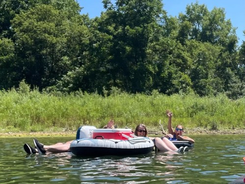 Two people relax on inflatable rafts in a calm lake, waving and enjoying a sunny day surrounded by greenery.