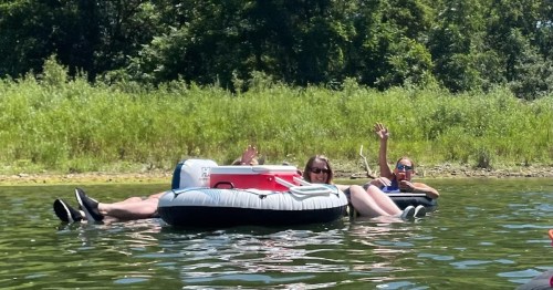 Two people relax on inflatable tubes in a calm lake, waving and enjoying a sunny day surrounded by greenery.