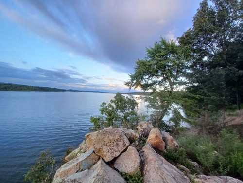 A serene lakeside view with rocks in the foreground, trees on the shore, and a cloudy sky reflecting on the water.