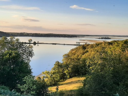 A serene landscape featuring a calm lake surrounded by trees and rolling hills under a clear sky at sunset.