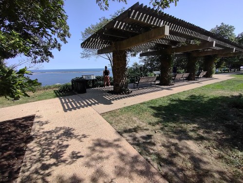 A shaded pergola with benches overlooks a scenic waterfront, surrounded by greenery and a clear blue sky.