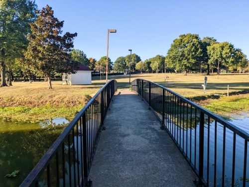 A concrete bridge over water, leading into a park with trees and a clear blue sky in the background.