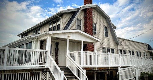 A large, two-story house with a white railing and a brick chimney, set against a cloudy sky.