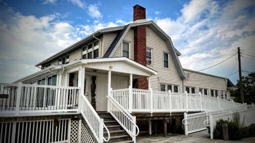 A large, two-story house with a sloped roof, white railings, and a brick chimney, set against a cloudy sky.